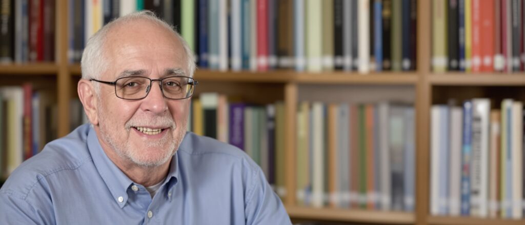 Smiling older man with glasses in front of a bookshelf, representing experienced PhD consultants providing dissertation writing support.